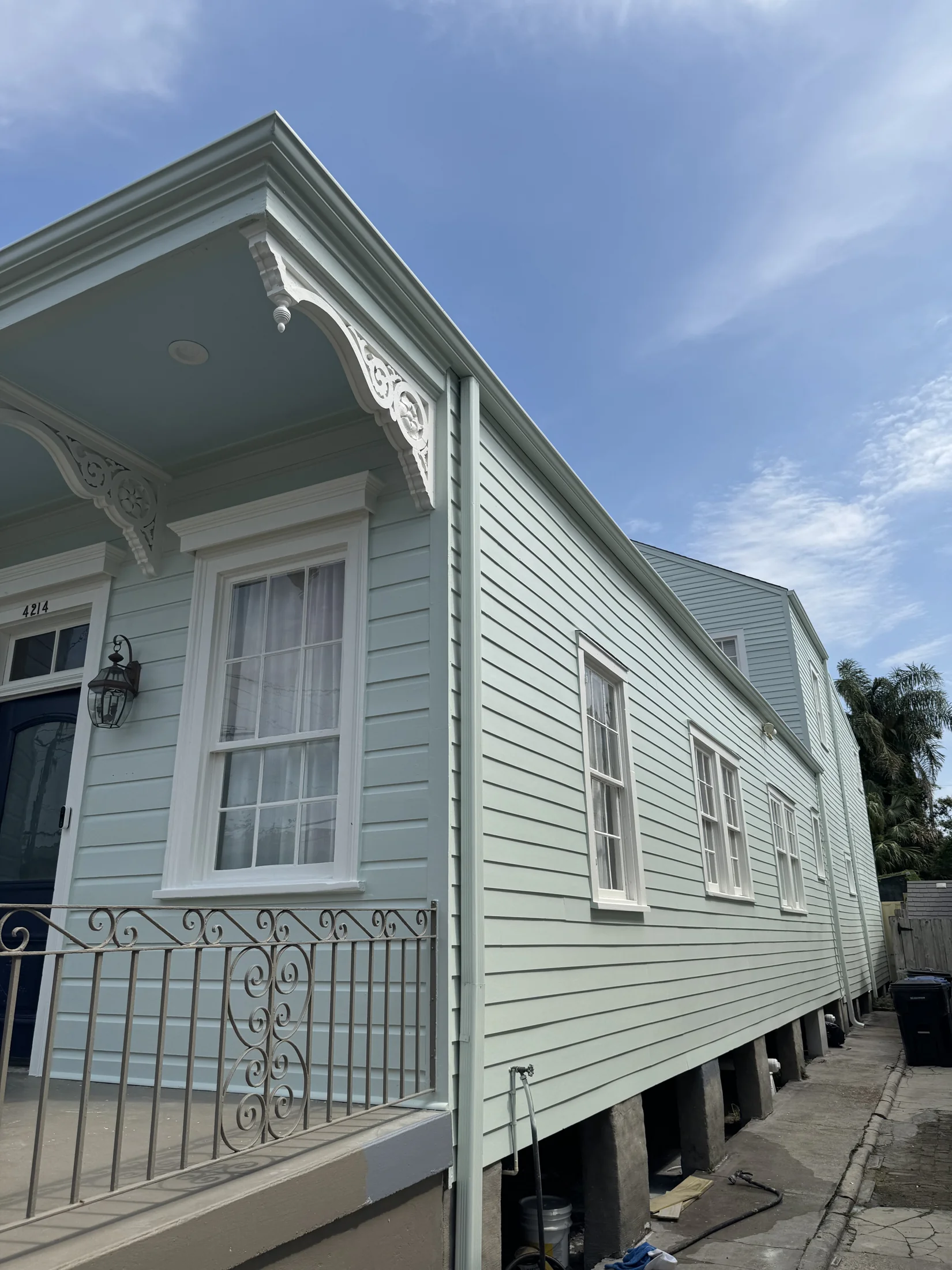 Freshly painted light blue New Orleans shotgun double with white trim, decorative eave brackets, and ornamental iron porch railing